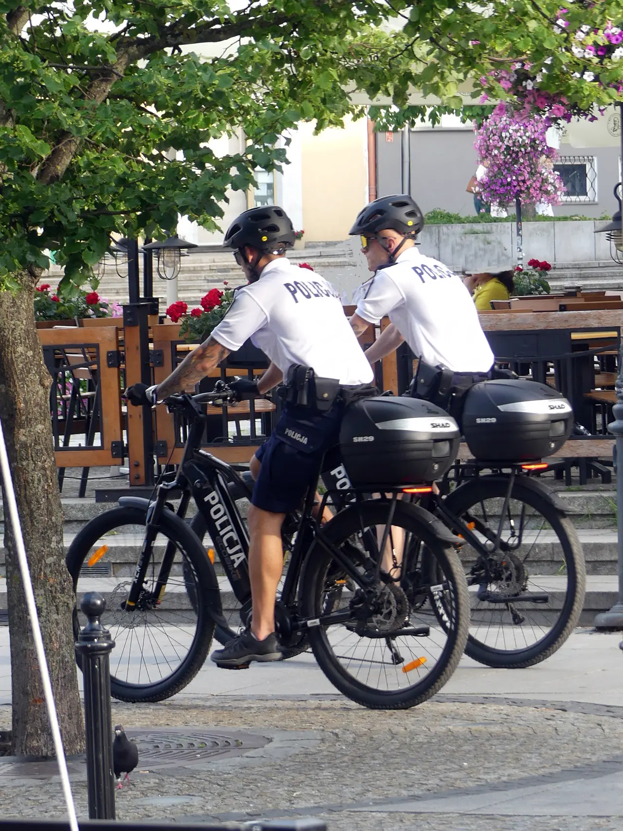 Zwei Polizisten mit Helm auf Dienstfahrrädern mit der Aufschrift Policja, von hinten.