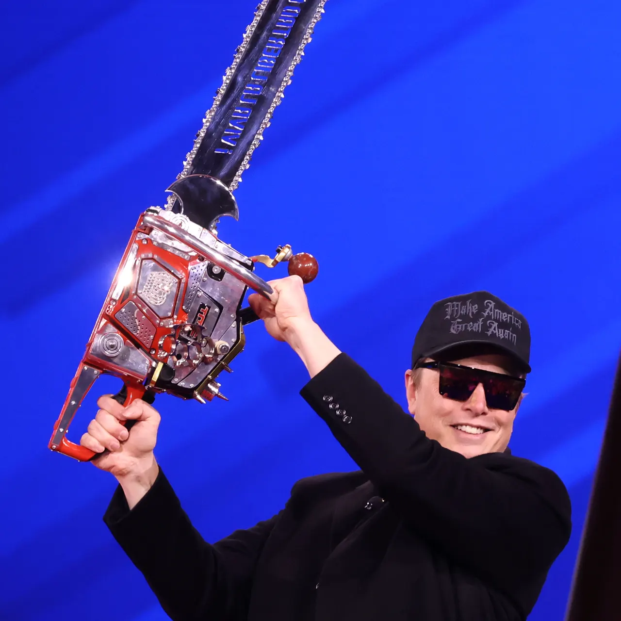 Elon Musk mit Sonnenbrille und MAGA-Kappe hält vor blauem Hintergrund eine Motorsäge hoch.