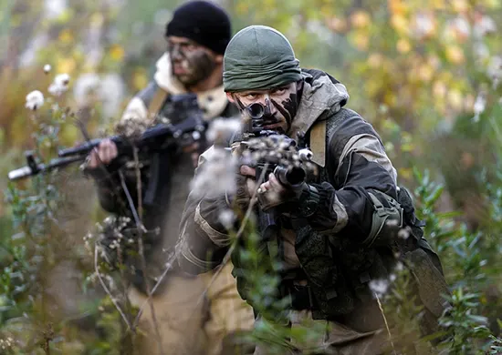Zwei bewaffnete Soldaten mit Tarnkleidung und Gesichtstarnung zwischen Bäumen.